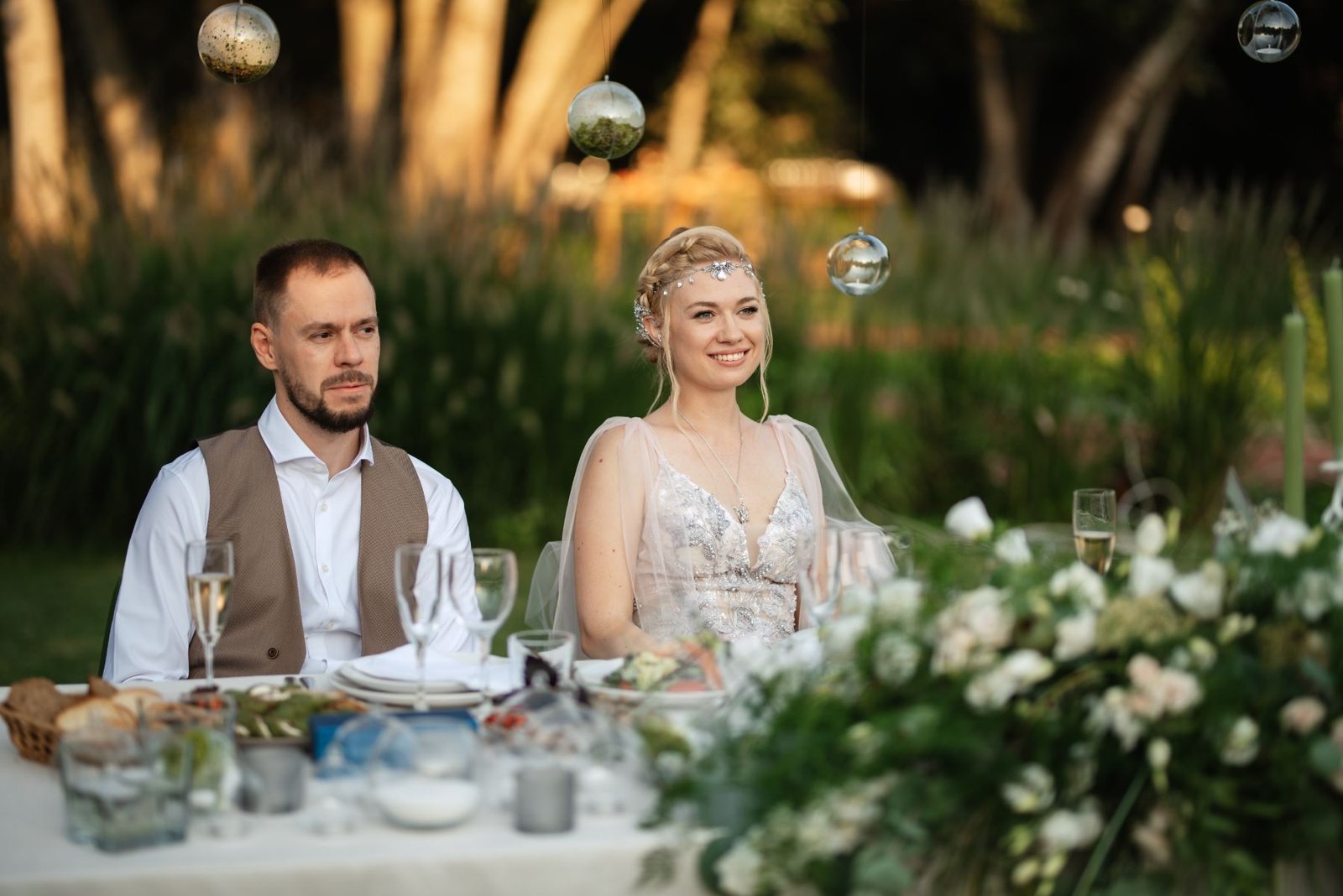 Bride and groom seated at a candlelit outdoor table with white flowers and hanging glass baubles