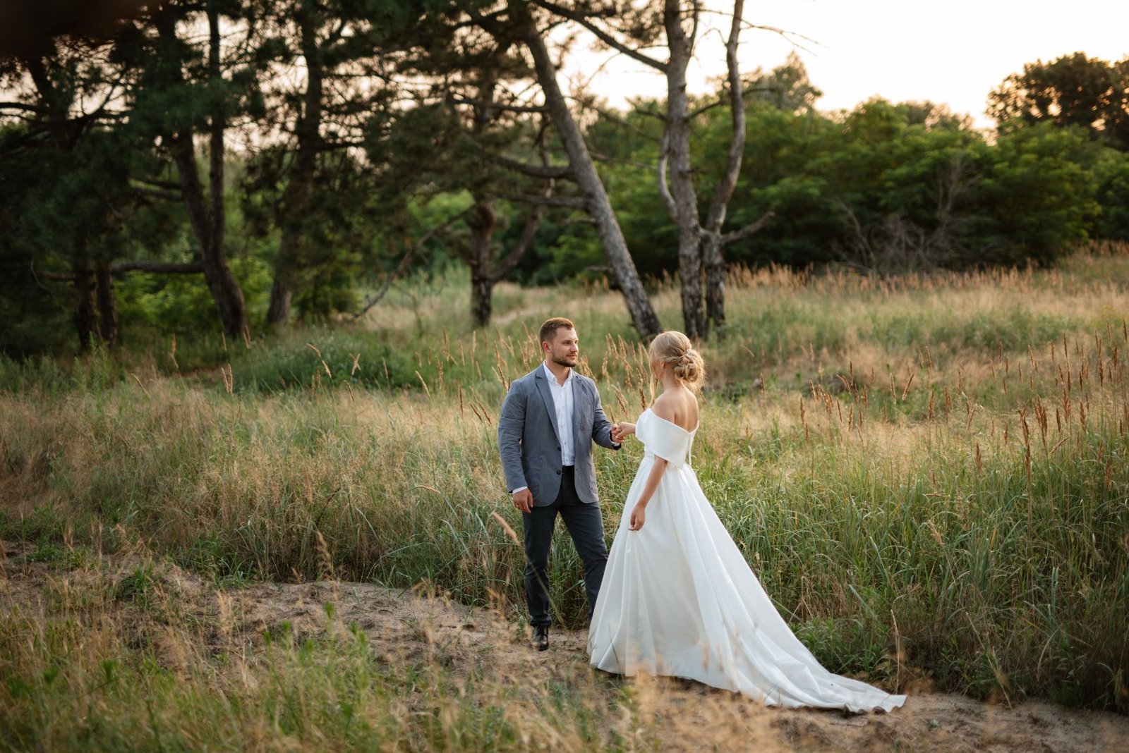 Couple holding hands on a sandy path through long grass beneath pine trees
